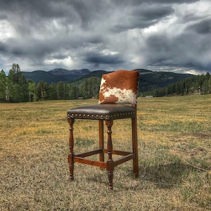 Puede incluir: Un taburete de bar de piel de vaca marr&oacute;n y blanca con un asiento de cuero marr&oacute;n oscuro y un marco de madera. El taburete se coloca sobre un fondo de campo de hierba y una cordillera en la distancia.