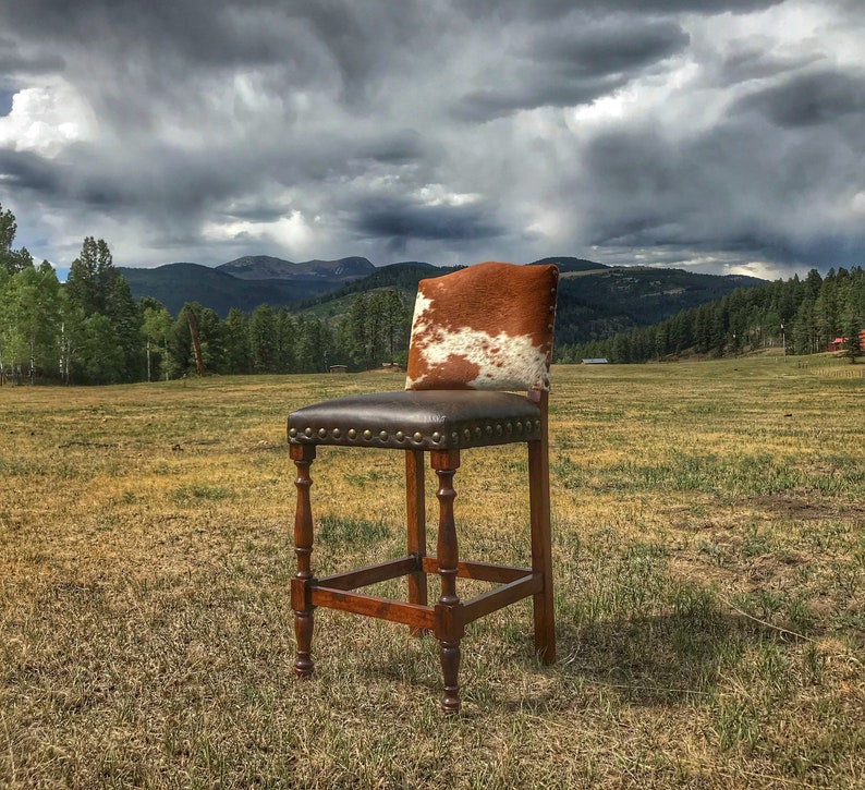 Puede incluir: Un taburete de bar de piel de vaca marr&oacute;n y blanca con un asiento de cuero marr&oacute;n oscuro y un marco de madera. El taburete se coloca sobre un fondo de campo de hierba y una cordillera en la distancia.