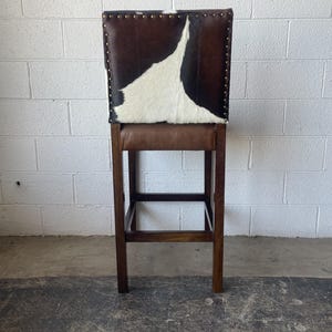 May include: A brown and white cowhide bar stool with a wooden frame. The stool has a rectangular seat and a footrest.