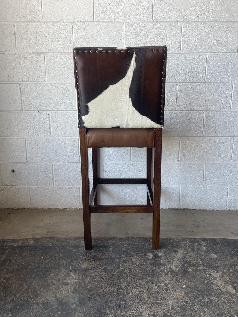 May include: A brown and white cowhide bar stool with a wooden frame. The stool has a rectangular seat and a footrest.