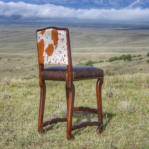 May include: A brown wooden bar stool with a cowhide seat and back. The stool is set against a backdrop of a grassy field and mountains.