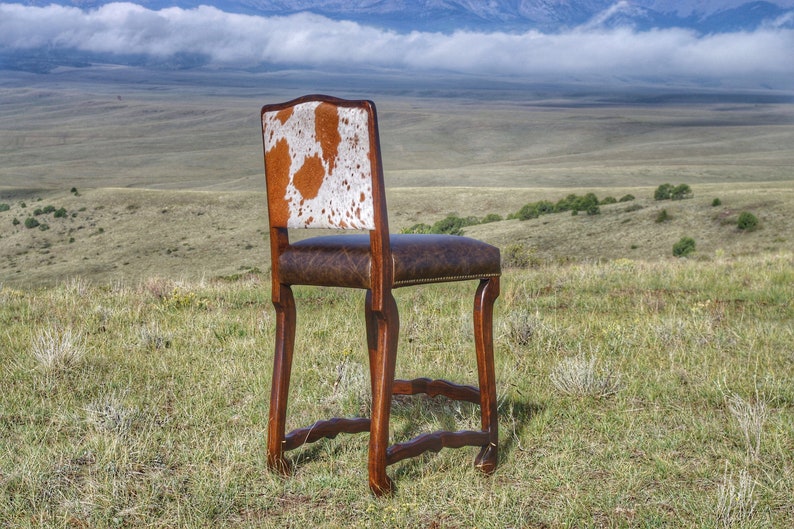 May include: A brown wooden bar stool with a cowhide seat and back. The stool is set against a backdrop of a grassy field and mountains.