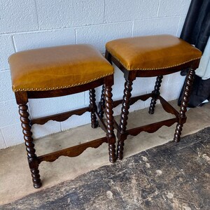 K&ouml;nnte beinhalten: Zwei braune Barhocker aus Holz mit einem braunen Ledersitz. Die Hocker haben ein gedrehtes Design an den Beinen.