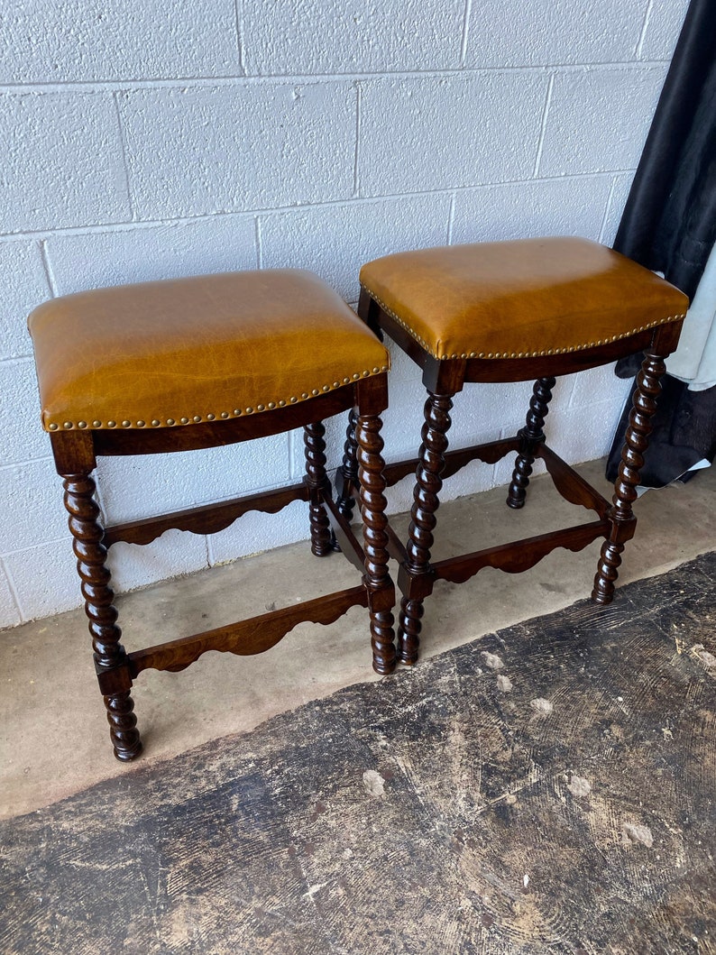 K&ouml;nnte beinhalten: Zwei braune Barhocker aus Holz mit einem braunen Ledersitz. Die Hocker haben ein gedrehtes Design an den Beinen.