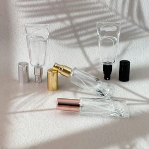 Könnte beinhalten: Vier durchsichtige Glassprühflaschen mit verschiedenen Verschlüssen. Zwei Flaschen stehen aufrecht mit silbernen und schwarzen Sprühköpfen, während zwei mit goldenen und roségoldenen Kappen flach liegen. Die Flaschen sind leer und zylindrisch.