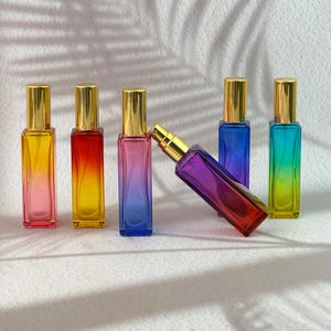 Könnte beinhalten: Eine Sammlung von sechs rechteckigen Glasparfümflaschen mit goldfarbenen Sprühkappen. Fünf Flaschen stehen aufrecht und zeigen lebendige Farbverläufe, darunter Gelb-zu-Rosa, Rot-zu-Gelb, Blau-zu-Violett, Blau sowie Grün-zu-Gelb.