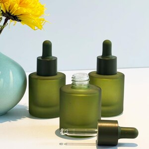 Puede incluir: Tres frascos cuentagotas de vidrio verde oliva con tapas y cuentagotas verde oscuro. Una botella está abierta, con el cuentagotas al lado. Una flor amarilla y un jarrón azul están en el fondo. Las botellas están sobre una superficie blanca.