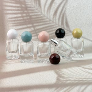 Puede incluir: Botellas de perfume de vidrio transparente con tapas esféricas de colores blanco, turquesa, rosa, negro y amarillo. Una botella está tumbada de lado y una esfera roja oscura está delante. Las botellas están sobre una superficie blanca.