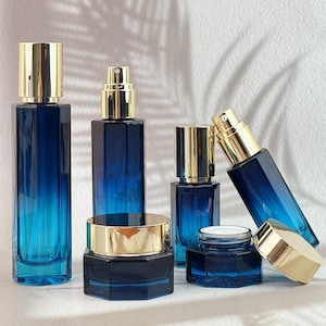 May include: An assortment of blue glass cosmetic bottles and jars with gold-colored lids. The bottles vary in shape and size, including a tall, rectangular bottle, a square bottle with a spray nozzle, and a small hexagonal jar. The blue color fades from dark to light.