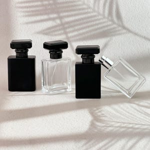 Puede incluir: Cuatro frascos de perfume rectangulares con tapones negros. Dos frascos son de vidrio negro opaco y dos de vidrio transparente. Los frascos transparentes tienen boquillas plateadas. Están alineados y uno de ellos está inclinado.