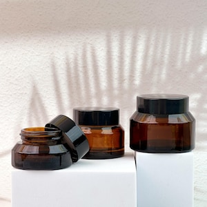 May include: Three amber glass jars with black lids are displayed on a white surface. One jar has its lid off to the side. The jars are cylindrical with a wide base and a short, rounded body. The background is a textured white wall with shadows.