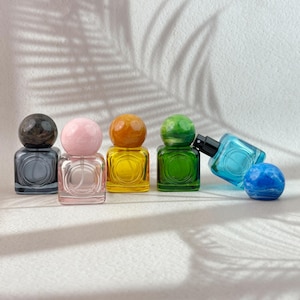 Puede incluir: Una colección de pequeños frascos de perfume coloridos con tapones esféricos. Los frascos son cuadrados y vienen en varios colores, incluyendo gris, rosa, amarillo, verde y azul. Los tapones también son de diferentes colores, dispuestos sobre una superficie blanca.