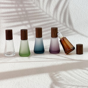 May include: A set of five small glass bottles with wooden caps. Four bottles have a tapered shape with frosted finishes in clear, green, blue, and purple. One amber glass bottle is tilted, showing a metal roller ball top with a separate wooden cap.