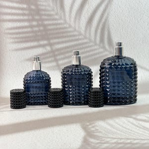 Op de afbeelding: Drie donkerblauwe glazen parfumflacons met zilveren verstuivers en zwarte getextureerde doppen. De flacons hebben een geometrisch patroon. De doppen zijn cilindrisch met een gestippelde textuur. De flacons hebben verschillende maten.