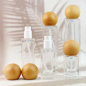 Peut inclure: Bouteilles en verre transparent avec des pompes blanches et des couvercles sphériques en bois. Les bouteilles sont rectangulaires avec un design incurvé. Les couvercles en bois sont brun clair. Les bouteilles sont disposées sur une surface blanche.