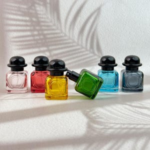 May include: A selection of miniature perfume bottles in various colours. Each bottle has a square design and a black bowler hat-shaped cap. The bottles contain liquids in shades of pink, red, yellow, green, blue, and grey.