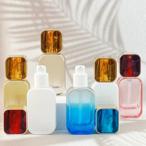 May include: An assortment of glass bottles with white pump tops and square, jewel-toned caps. The bottles are in various colors, including clear, white, blue, and pink. The caps are gold, amber, red, and blue. The bottles are arranged on a white surface.