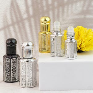 Puede incluir: Una variedad de frascos de perfume decorativos en oro, plata y negro. Los frascos tienen patrones grabados ornamentados y están dispuestos sobre una superficie blanca. Una flor amarilla brillante está en el fondo.