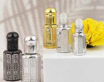 May include: An assortment of decorative perfume bottles in gold, silver, and black. The bottles have ornate, engraved patterns and are arranged on a white surface. A bright yellow flower is in the background.
