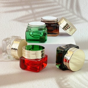 May include: A selection of small, square glass jars in various colours, including clear, green, red, and brown. Each jar has a gold-coloured lid. The jars are arranged on a white surface, with soft shadows in the background. The jars are empty.