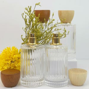 May include: Clear glass perfume bottles with gold sprayers and wooden caps. The bottles have a ribbed design and are displayed with yellow flowers and dried greenery. The wooden caps are in shades of light and dark brown.