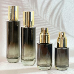 May include: Four glass spray bottles with gold-colored caps. The bottles have a gradient effect, transitioning from a dark gray at the base to a lighter shade towards the top. The bottles are of varying heights.
