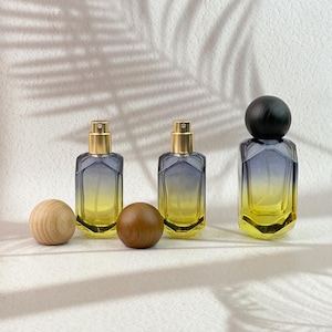 May include: Three perfume bottles with gold sprayers and a gradient design of yellow and blue. Two wooden spheres and a black wooden sphere are next to the bottles. The bottles are on a white surface.