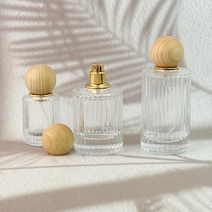 Puede incluir: Tres frascos de perfume de vidrio transparente con texturas acanaladas verticales y tapas esféricas de madera clara. Un frasco se muestra sin tapa, revelando un atomizador dorado. Los frascos varían en altura y están dispuestos en grupo.