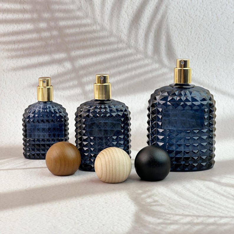 Three navy blue diamond cut glass spray bottles with gold spray pumps, dark/light/black wood caps placed in front on white textured background.