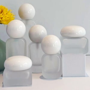 Op de afbeelding: Een verzameling witte keramische bollen en containers van matglas. De bollen staan bovenop de containers, wat een minimalistische en moderne esthetiek creëert. De opstelling omvat verschillende vormen en maten en biedt een strakke en elegante presentatie.
