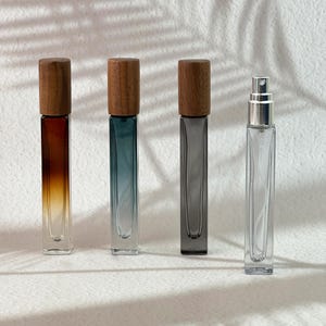 Op de afbeelding: Vier hoge, rechthoekige glazen parfumflesjes op een rij. Drie flesjes hebben houten doppen en glas met een kleurverloop in amber, blauwgroen en grijs. Het vierde flesje is van helder glas met een zilverkleurige metalen verstuiver.