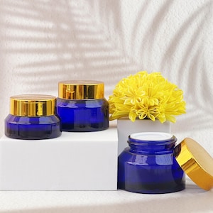 Könnte beinhalten: Drei kobaltblaue Glasbehälter mit goldfarbenen Deckeln. Die Behälter sind unterschiedlich groß und stehen auf einer weißen Oberfläche. Im Hintergrund befindet sich ein gelbes Blumenarrangement. Die Behälter sind wahrscheinlich für Kosmetika oder Hautpflegeprodukte.