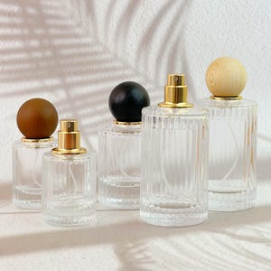 May include: Five clear glass perfume bottles with gold sprayers and wooden or black spherical stoppers. The bottles have a ribbed design and are arranged on a white surface. The stoppers are made of light and dark wood, and black material.