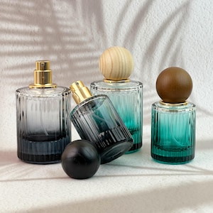 May include: Four glass perfume bottles with gold sprayers and wooden or black stoppers. The bottles have a ribbed design and gradient colors, ranging from clear to teal and dark blue. The bottles are arranged on a white surface.