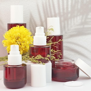 Könnte beinhalten: Eine Sammlung roter Kosmetikbehälter aus Glas mit weißen Pumpspendern und Deckeln. Die Behälter sind mit einer gelben Blume und Zweigen aus Grün angeordnet. Der Hintergrund ist weiß mit Schatten.
