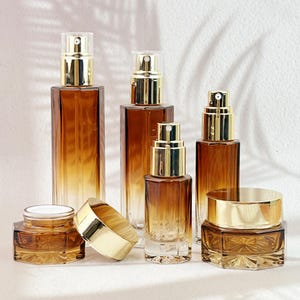 Op de afbeelding: Een assortiment amberkleurige glazen cosmeticaflessen en potten met goudkleurige doppen en deksels. De flessen variëren in grootte en vorm, waaronder spuitflessen en een pot met een schroefdeksel. De opstelling suggereert een huidverzorgings- of schoonheidsproductlijn.