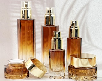 May include: An assortment of amber-colored glass cosmetic bottles and jars with gold-colored caps and lids. The bottles vary in size and shape, including spray bottles and a jar with a screw-on lid. The arrangement suggests a skincare or beauty product line.