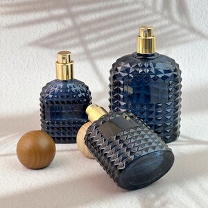 Three navy blue diamond cut glass spray bottles, one tilted to show spray pump, with dark/light wood caps on white textured background.