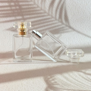 Puede incluir: Frascos de perfume de vidrio transparente con formas rectangulares y tapones de cristal. Un frasco tiene un pulverizador dorado, el otro plateado. Los frascos están vacíos y los tapones están sueltos.