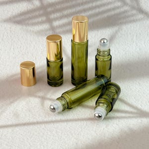May include: A collection of olive green glass roller bottles with shiny gold caps. Two bottles stand upright with caps on, one stands open with a metal roller ball exposed, and two bottles lie horizontally, one showing the metal roller ball mechanism.
