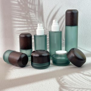 May include: A collection of frosted sage green glass cosmetic containers featuring dark brown wooden caps. The set includes various sizes of cylindrical bottles with white pump dispensers and round jars, some with lids removed to reveal the interior.