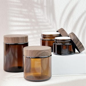 May include: Four amber glass jars with wooden lids. The jars are cylindrical and vary in size, with the lids featuring a wood grain pattern. The jars are arranged on a white surface, with a soft, neutral background.