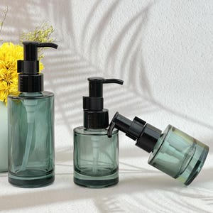 Puede incluir: Tres botellas de vidrio transparente con dispensadores de bomba negros. Las botellas tienen un tinte verde suave y son cilíndricas o redondeadas. Las botellas están dispuestas sobre una superficie blanca, con una sombra de hojas en el fondo.