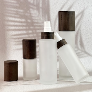 Puede incluir: Una colección de botellas de spray de vidrio esmerilado con tapas de madera marrón oscuro. Las botellas varían en tamaño y forma, con algunas de pie y una inclinada. El fondo es una superficie blanca lisa con sombras.