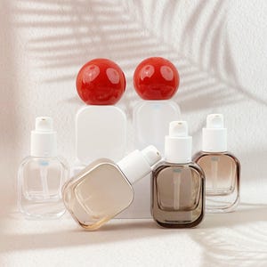 May include: Clear and amber glass pump bottles with white pumps. Two red spherical objects sit atop white square bases. The bottles are arranged on a white surface, suggesting a skincare or cosmetic product.