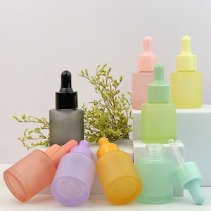 May include: A selection of pastel-coloured frosted glass dropper bottles. The bottles are in shades of pink, yellow, green, orange, and purple, with one featuring a black dropper. The bottles are arranged with decorative greenery.