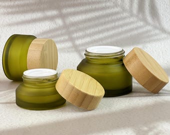 May include: Three round cosmetic jars made of frosted olive green glass with light-colored bamboo lids. Two jars are open with white inner liners visible, while one jar is closed. The jars have a low-profile, wide-mouth design.