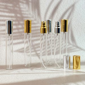 May include: A collection of clear glass perfume or essential oil spray bottles with silver and gold-colored caps. The bottles are rectangular and have a fine mist sprayer. The caps are cylindrical and sit atop the bottles. The background is a textured white surface.