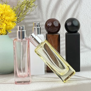 May include: A selection of perfume bottles in different colours and designs. The bottles are rectangular with silver atomisers. Colours include clear, pink, white, brown, and black. Some bottles have spherical stoppers.
