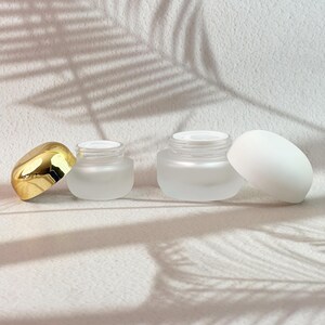 Op de afbeelding: Twee cosmetische potjes van matglas met witte deksels. Eén potje heeft een gouden deksel ernaast. Het andere potje heeft een witte deksel ernaast. De potjes staan op een wit oppervlak met de schaduw van een blad.